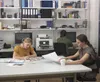 Three people working in an office environment with shelves of books and office supplies in the background. Náhled obrázku produktu
