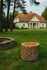 A wooden pouffe sits on a grassy lawn in front of a white house with a red roof. A stone fire pit is visible to the left. Podgląd zdjęcia produktu