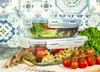 Two glass food storage containers filled with food, placed on a checkered cloth with breadsticks and tomatoes in the foreground. Produktbild-Vorschau