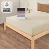 A bed with a cream-colored fitted sheet is shown, with a stack of folded sheets in various colors on top of it. A small table with a vase and a book is in the background. Produktbild-Vorschau