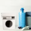 A white storage container designed to look like a washing machine, placed on a white wooden surface. Next to it are laundry detergent, laundry pods, and a basket of blue towels. Náhled obrázku produktu
