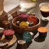 A collection of coffee capsules arranged around a wooden bowl, with a cup of espresso and a coffee machine in the background. Produktbild-Vorschau
