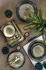 A table setting with dark blue and beige ceramic plates, bowls, cutlery, and a glass, arranged with a fern plant and candles. Produktbild-Vorschau