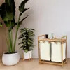 A bamboo laundry basket with two beige fabric bags is placed next to two large potted plants in a room with light-colored walls and wooden flooring. Anteprima dell’immagine prodotto