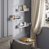 Three white shelves are mounted on a light gray wall, displaying decorative items. A sofa and a small table with a cup of coffee are in the foreground, and a window with curtains is on the right. Produktbild-Vorschau