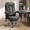 A black ergonomic office chair with a footrest is shown in a room with a desk and shelves in the background. Náhled obrázku produktu
