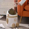 A person is shown throwing crumpled paper into a cute, astronaut-shaped trash can. The trash can is white with tan accents and has the number '10' on its side. It is placed on a carpeted floor next to a dresser and an orange armchair. Produktbild-Vorschau