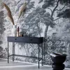 A black console table with fluted drawers and metal legs is placed against a black and white forest-themed wallpaper. Decorative items include a white vase with dried grasses, and a stack of books on a black stool. Produktbild-Vorschau