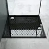 A black square shower tray with a drain and textured surface, placed on a concrete floor inside a shower enclosure. Produktbild-Vorschau