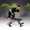 A man is performing a dumbbell row exercise on a black adjustable weight bench. The word "Grind" is displayed in large, glowing yellow letters in the background. Produktbild-Vorschau