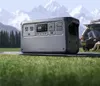 A portable power station is set up on a mat in a grassy field with mountains and trees in the background. A car is parked nearby, and two people are sitting in the back. Náhled obrázku produktu