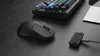 A black wireless mouse and a small black accessory are placed on a dark, textured surface next to a portion of a keyboard. Náhled obrázku produktu