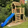 A wooden play structure with a blue slide, climbing wall, and ladder is set up in a grassy backyard with trees and bushes in the background. Náhľad obrázku produktu