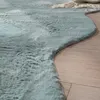A close-up view of a light blue faux fur rug with a wavy edge, lying on a light wood floor. A small section of a beige blanket with fringe is visible at the top right. Anteprima dell’immagine prodotto