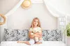 A young girl sits on a bed holding a teddy bear, with pillows and a canopy in the background. Produktbild-Vorschau