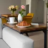 A close-up of a C-shaped side table placed on the armrest of a sofa, with a croissant and coffee cup on it, and a vase of flowers and a yellow chair in the background. Produktbild-Vorschau
