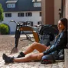 A woman sits on a cobblestone ground next to a vintage-style electric bicycle, leaning against a building. Náhled obrázku produktu