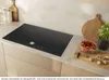 A modern induction cooktop is shown in a kitchen setting, with some food items and kitchenware on the counter. Voir les images du produit