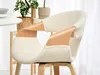 A close-up view of a cream-colored armchair with wooden accents, placed in a well-lit room with plants and a table in the background. Produktbild-Vorschau