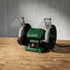 A green and gray bench grinder sits on a wooden surface against a concrete wall. Produktbild-Vorschau