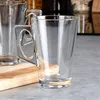 Two clear glass mugs with square handles are placed on a wooden board, with a blurred background of kitchen shelves. Voir les images du produit