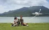 Two young women and a dog sitting on grass by a lake with mountains in the background, and a drone flying in the sky. Anteprima dell’immagine prodotto