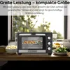 A mini oven is shown on a wooden surface with food items around it, with a blurred outdoor scene visible through a window in the background. Produktbild-Vorschau