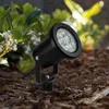 A black LED garden spotlight is planted in the ground next to some green foliage and white flowers. Náhled obrázku produktu