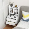 A white bedside table with drawers and shelves is placed next to a bed with a gray headboard and a colorful pillow. Podgląd zdjęcia produktu
