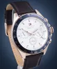A close-up studio shot of a Tommy Hilfiger watch with a white dial, rose gold casing, and a brown leather strap, set against a dark blue background. Anteprima dell’immagine prodotto