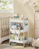 A white rolling cart filled with baby care items is placed next to a crib in a nursery. Produktbild-Vorschau