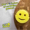A yellow Scrub Daddy sponge with a smiley face is placed on a wooden cutting board next to a white towel. Text overlays highlight its scrubbing power. Produktbild-Vorschau