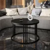 A round black coffee table with a vase of dried flowers and a cup of coffee sits in front of a curved gray sofa in a modern living room. Produktbild-Vorschau