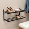 A black metal shoe rack mounted on a light gray wall, displaying several pairs of women's shoes and a folded stack of towels. Produktbild-Vorschau