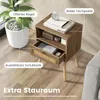 A nightstand with a drawer and open shelf is shown in a room setting with a plant and part of a sofa. Podgląd zdjęcia produktu