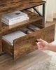 A hand is reaching to open a drawer on a rustic brown TV stand with black metal accents. Books and a remote control are visible inside the drawers. Náhled obrázku produktu