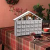 A Christmas advent calendar decorated with miniature houses and trees, set on a table with a plaid tablecloth. Voir les images du produit