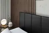 A close-up view of a black upholstered headboard of a bed, with a bedside table and lamp visible on the left. Produktbild-Vorschau
