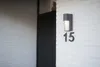 A modern outdoor wall light is mounted on a white, horizontally paneled wall next to a dark gray door. The house number '15' is displayed below the light. Anteprima dell’immagine prodotto