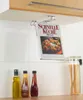 A kitchen scene with a cookbook holder mounted under a cabinet, holding a cookbook. Several bottles of oil and sauce are on the counter below, along with a jar of pasta. Produktbild-Vorschau