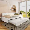 A white daybed with a pull-out trundle bed is set up in a bright, airy room with a large window overlooking a green landscape. The room has a modern, minimalist aesthetic with arched wall niches and shelves. Náhled obrázku produktu
