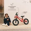 A young child sits next to a red balance bike against a textured wall. Produktbild-Vorschau