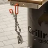 A pair of metal tongs with orange handles hangs from a hook attached to a grill. Podgląd zdjęcia produktu