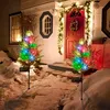Two solar-powered cypress trees with colorful LED lights are placed in the snow in front of a house with a red door decorated for Christmas. Anteprima dell’immagine prodotto