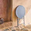 A round, gray-blue bistro table is folded and leaning against a textured, tan wall. The table is on a stone patio. To the left, there's a wooden door and a dark lantern. Anteprima dell’immagine prodotto