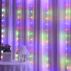 A table with a lamp and a plant in front of sheer curtains with colorful string lights. Produktbild-Vorschau