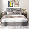 A bedroom scene featuring a bed with a gray upholstered headboard and frame, dressed with bedding and pillows. Two wooden nightstands are placed on either side of the bed, each with a lamp and decorative items. Artwork hangs on the wall above the bed, and  Podgląd zdjęcia produktu