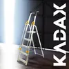 A silver aluminum ladder with yellow accents stands in a dimly lit industrial-looking space with a dark corrugated metal wall in the background and a bright light source on the left. Podgląd zdjęcia produktu