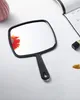A black hand mirror is placed on a white surface, with a pink vase and a book in the background. Produktbild-Vorschau