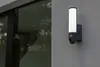 A wall-mounted outdoor light fixture with a built-in camera is shown on a gray wall next to a large window. Produktbild-Vorschau
